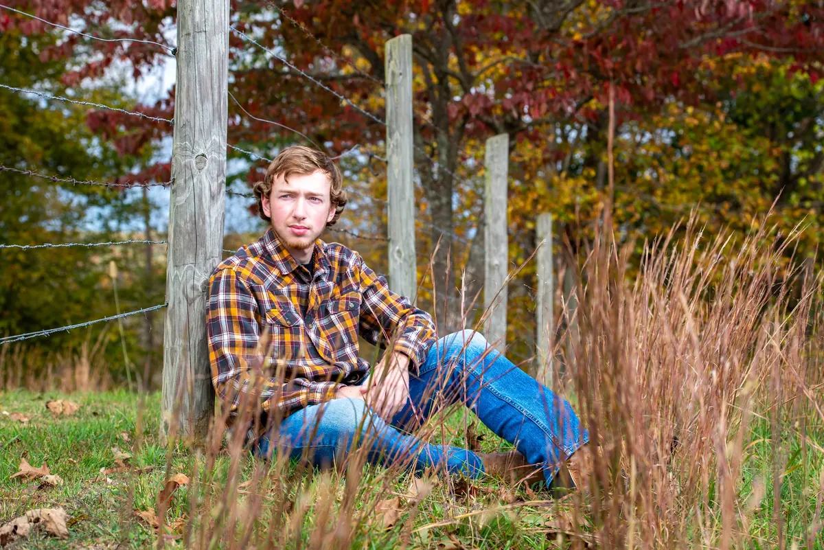 Senior in a plaid shirt seated beside a fence in autumn grass
