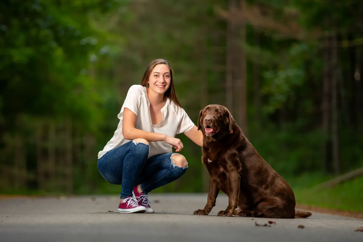 Senior crouching beside a brown dog on a wooded road